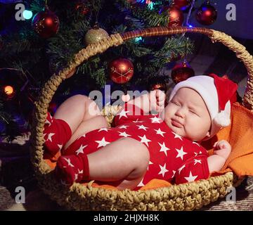 Neugeborenes Baby schläft in einem Weihnachtsmannhut unter einem Weihnachtsbaum in einem Korb. Speicherplatz kopieren. Stockfoto