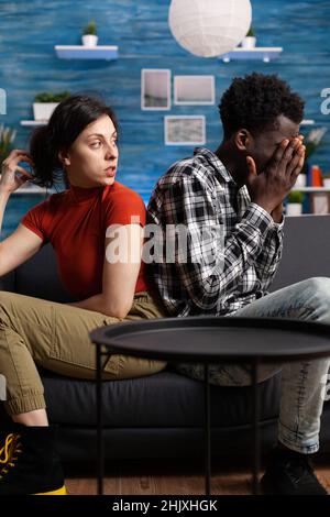Unzufriedenes interrassisches Paar, das in Stille auf dem Sofa sitzt, während kaukasische Frau wütend über die Schulter zum weinenden afroamerikanischen Mann schaut. Erwachsene, die Eheprobleme haben, während der Mann besorgt ist Stockfoto