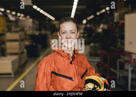 Lächelnde junge Arbeiterin in orangefarbener Schutzkleidung in der Fabrik Stockfoto