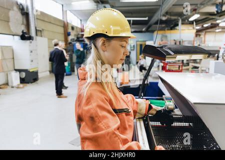 Seitenansicht der weiblichen Angestellten in Hardhut, die Maschinen in der Fabrik benutzen Stockfoto