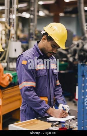 Männlicher Arbeiter mit blauem Kragen, der in Notizblock schreibt, während er in der Industrie arbeitet Stockfoto