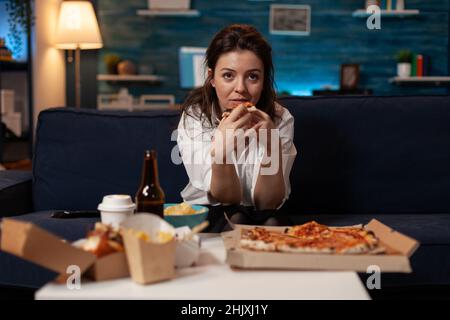 Person, die auf der Couch im Wohnzimmer sitzt und eine Scheibe Pizza zum Mitnehmen hat, während sie am Tisch eine Sitcom-Fernsehsendung mit Schnellimbiss zum Mitnehmen anschaut. Frau nach der Büroarbeit Essen zum Mitnehmen tv-Abendessen. Stockfoto