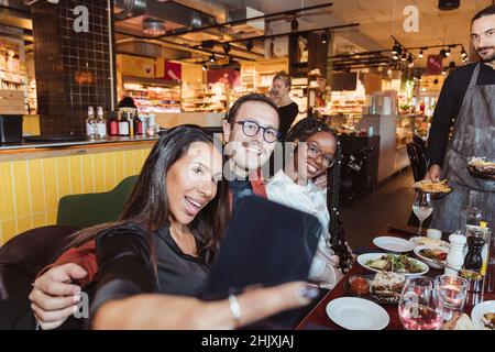 Fröhliche Frau, die mit männlichen und weiblichen Freunden im Restaurant Selfie über das Smartphone nimmt Stockfoto
