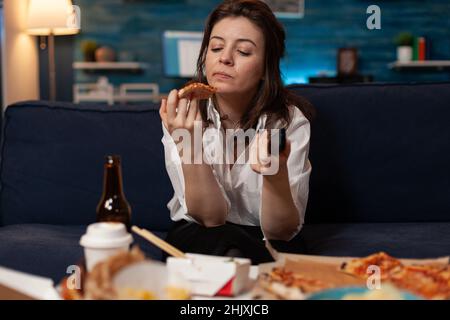 Frau schaut auf ein Stück Pizza, die nach Hause geliefert wird, während sie die Fernschaltkanäle auf dem Fernseher vor dem Couchtisch mit einem Menü zum Mitnehmen hält. Person, die Fast-Food-Sender durchsucht. Stockfoto