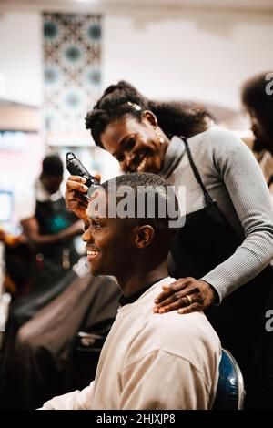 Lächelnder weiblicher Barbier, der die Haare eines männlichen Kunden mit einem elektrischen Rasiermesser im Salon schneidet Stockfoto