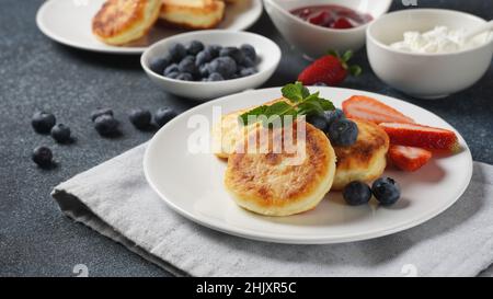 Quark-Fritten oder Syrniki oder Pfannkuchen mit frischen Blaubeeren, Erdbeeren und Erdbeermarmelade. Leckeres Dessert oder süßes Frühstück. Russisch, Ukr Stockfoto