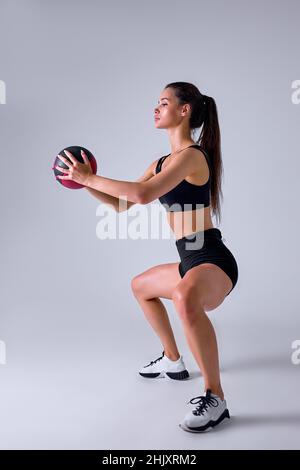 Seitenansicht Porträt von Slim Caucasian Cross fit Frau mit Fitness-Ball hockt, isoliert im Studio. Fit Lady trainiert mit Medizinball. Koac Stockfoto
