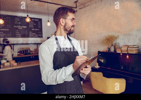 Fröhlicher männlicher Arbeiter, der im Café auf die Zwischenablage schrieb Stockfoto