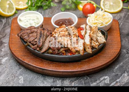 Fleisch- und Hühnerfajitas auf schwarzem Steinteller mit Saucen Stockfoto