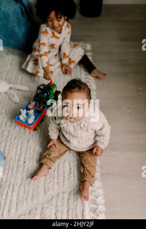 Männliches Kleinkind mit Schnuller sitzt auf dem Teppich, während die Schwester im Hintergrund zu Hause sitzt Stockfoto