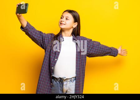 Kleines Kind gir über gelbem Hintergrund hält modernes Mobiltelefon und lässt Videoanruf in Hello-Geste Palme. Stockfoto