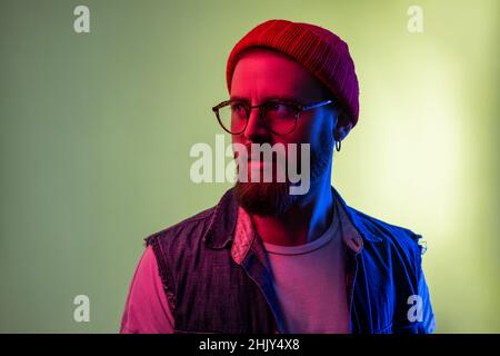 Porträt eines jungen trendigen Hipster-Mannes in einer Brille, die posiert und mit einem ernsthaft durchdachten Gesicht wegschaut, mit Beanie-Hut und Denim-Weste. Innenaufnahmen im Studio, isoliert auf farbigem Neonlicht-Hintergrund. Stockfoto