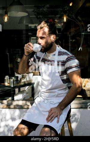 Junger männlicher Besitzer trinkt Kaffee, während er draußen im Café sitzt Stockfoto