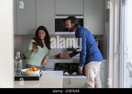Lächelnde Frau, die in der Küche ein Smartphone mit einem Mann teilt Stockfoto