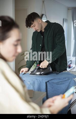 Junger Mann bügelt Hemd, während Freundin Smartphone im Wohnzimmer benutzt Stockfoto