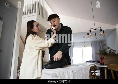 Frau, die Smartphone am Ohr des Freundes hält, während sie zu Hause Kleidung bügelt Stockfoto