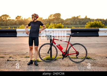Sportlicher Profi-Radfahrer in schwarzem Helm, Schutzbrille und aktiver Kleidung haben Ruhe nach dynamisch Fahrrad fahren. Mann nach Wettkämpfen und Stockfoto