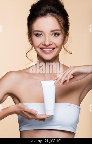 Lächelnde Brünette Frau in weiß oben hält kosmetische Lotion isoliert auf beige Stockfoto