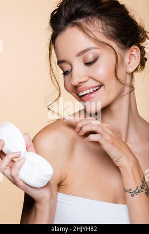 Fröhliche Frau, die kosmetische Creme auf der Schulter auf beige isoliert Stockfoto