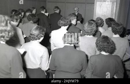 1950s, historisch, eine christliche Gemeinde. Nmen und Frauen stehen eng zusammen in einem kleinen Raum, lesen aus Bibeln. Ein männlicher Pastor in Anzug und Krawatte steht am anderen Ende des Raumes und blickt auf sein Publikum, die USA. Stockfoto