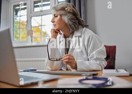 Gestresst Und Überarbeitet Reife Weibliche Doctor Trägt Weißen Mantel Am Schreibtisch In Ärzte Büro Stockfoto