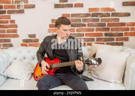 Junger Mann spielt E-Gitarre. Lernt Gitarre zu spielen. Interessantes Hobby. Weißes Sofa im Zimmer. Angenehme Erholung. Stockfoto