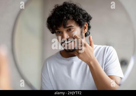 Happy Handsome indian Mann Anwendung Feuchtigkeitscreme, Pflege für die Haut unter den Augen und Blick auf runden Spiegel im Badezimmer Stockfoto