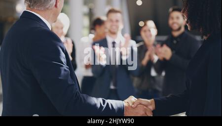 Harte Arbeit wird nie unerkannt. 4K Videoaufnahmen von zwei Geschäftsleuten, die sich in einem Büro die Hände schütteln, während Kollegen im Hintergrund applaudieren. Stockfoto