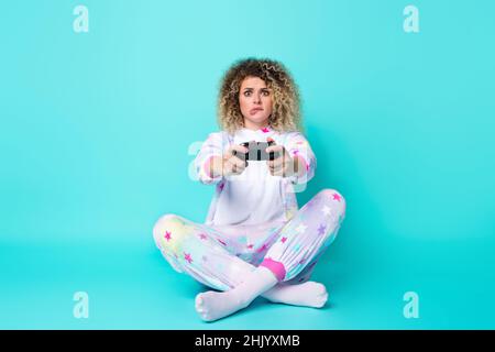 Full Lenght Foto von traurigen jungen lockigen Frisur Dame spielen Station Bite Lippe tragen Pyjama Socken isoliert auf blauem Hintergrund Stockfoto