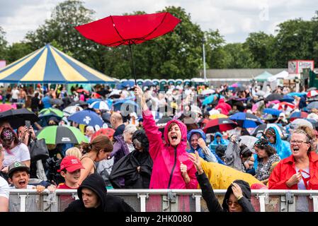 Fans bei einem großen Musikfestival in einem Park in Essex bei starkem Regen. Frau genießt die Show, obwohl ihr Regenschirm sich von innen nach außen dreht. Stoischer Spaß Stockfoto