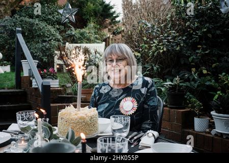 Frau saß mit einem Happy Birthday Abzeichen und Kuchen mit Wunderkerzen Stockfoto