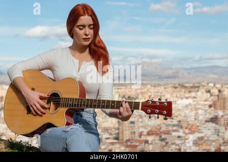 Rothaarige junge Frau spielt die Gitarre im Freien sitzend. Stockfoto