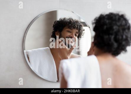 Hübscher indischer Mann, der im Badezimmer im Spiegel schaute und den Bart berührte und morgens die Schönheitsroutine genoss Stockfoto