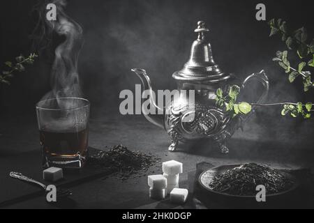 Traditionelle marokkanische Teekane mit einer dampfenden Tasse Tee, Zucker und Minze in rauchiger schwarzer Atmosphäre. Marokkanisches Teekonzept. Stockfoto