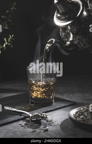 Traditionelle marokkanische Teekane gießt Tee in ein dampfendes Glas mit natürlichem Tee und Minze auf dunklem Hintergrund. Traditionelles marokkanisches Teekonzept. Stockfoto