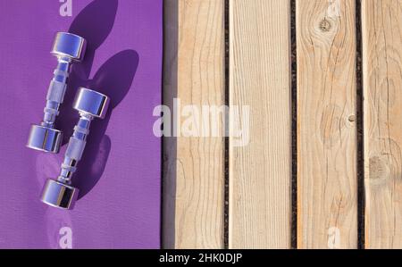 Violette Gummimatte mit Metallhanteln für das Fitnessstudio. Sport-Fitness-Accessoires draußen auf Holzboden an sonnigen Tagen. Stockfoto