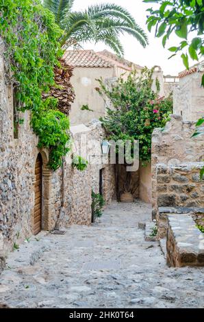 Schöne traditionelle mittelalterliche Straße auf der Insel Monemvasia, Peloponnes, Griechenland. Byzantinische Allee mit Steinhaus-Eingängen, Bäumen und Pflanzen. Stockfoto