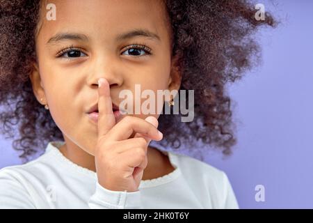 Entzückende schwarze kleine Mädchen hält Finger auf den Lippen Symbol der Stille Geste zu fragen, um Sei still. Stille oder geheimes Konzeptbild isoliert auf violettem Stu Stockfoto