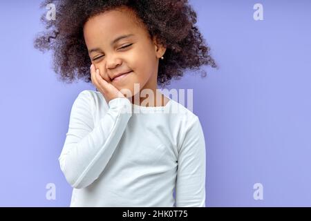 Happy girl Kid hält die Hände auf den Wangen, lächelt zufrieden, freundlich, mit geschlossenen Augen, hat Spaß mit Freunden zu plaudern, trägt lässige Kleidung, isoliert über pur Stockfoto