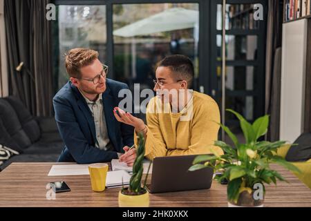 Lächelndes Paar verschiedener Geschäftsleute, die Schreibarbeiten machen und am Laptop arbeiten, während sie an einem Tisch im legeren Heimbüro sitzen. Stockfoto