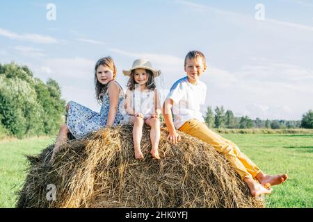 Porträt von drei Jungen und zwei Mädchen, die auf dem Heuhaufen auf dem Feld sitzen. Leichter, sonniger Tag. Fröhlich und genießen Konzept. Vorderansicht. Bäume und klarer Himmel auf Stockfoto