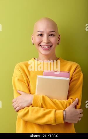 Porträt einer Frau mit kahlem Kopf, die Bücher in den Händen hält und lächelt, die Bildung genießt, studiert. Haarlose Dame in lässigem gelben Hemd posiert bei der Kamera Stockfoto