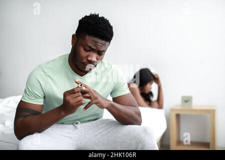 Unglücklicher, trauriger, tausendjähriger afroamerikanischer Kerl zieht den Ehering ab, beleidigte Dame, die auf dem weißen Bett im Schlafzimmer weinte Stockfoto