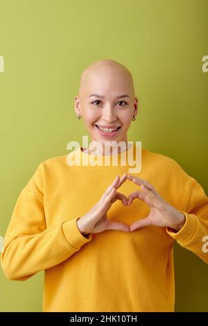 Kahlköpfige kaukasische Femalle in gelbem Hemd, die vor der Kamera posiert und das Herz aus den Händen zeigt, lächelnd, isoliert auf grünem Studiohintergrund. Menschliche Emotio Stockfoto