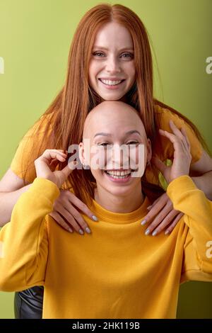 Zwei verschiedene europäische Rothaarige und haarlose Damenmodelle umarmen isoliert auf grünem Hintergrund, Porträt von jungen Frauen in lässigen gelben Hemden havi Stockfoto