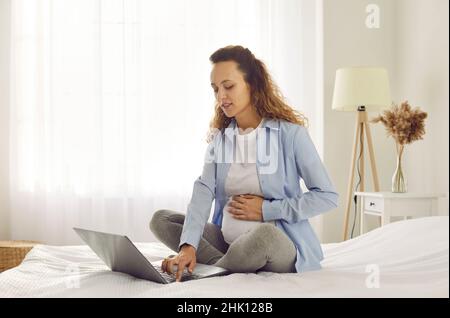 Schwangerin sitzt auf dem Bett im Schlafzimmer und mit dem Laptop auf der Suche nach Informationen über die Schwangerschaft. Stockfoto