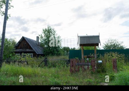Alte Holzbrunnen für Wasser mit einem Metalleimer in einem russischen Dorf neben einem einstöckigen Holzhaus in einem Dickicht aus grünem Gras und Sträuchern im Sommer. Stockfoto
