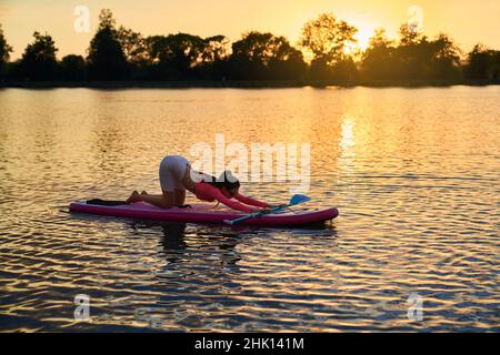 Schlanke junge Frau in activewear Stretching Körper auf supboard während schönen Sommer Sonnenuntergang. Konzept von Menschen, Ausgleich und Wasseraktivität. Stockfoto