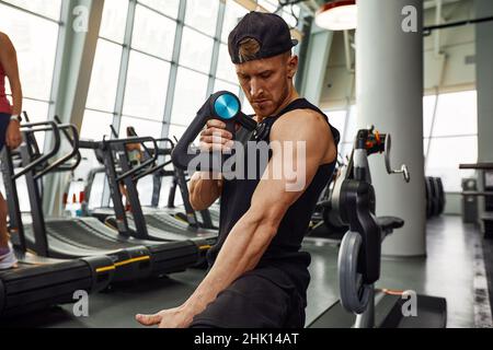 Ein junger Mann benutzt und genießt die Schlagmassagepistole, um sich zu entspannen und die Schmerzen im Körper und in den Muskeln im Fitnessstudio zu lindern. Stockfoto