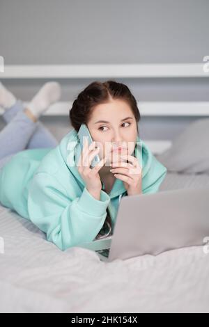 Fernunterricht Online-Bildung und Arbeit. Happy Brunette Frau Mädchen arbeiten Büro arbeiten entfernt von zu Hause in türkisfarbenen Hoodie und Jeans auf dem Bett. Telefon sprechen. Mit Computer Laptop Stockfoto